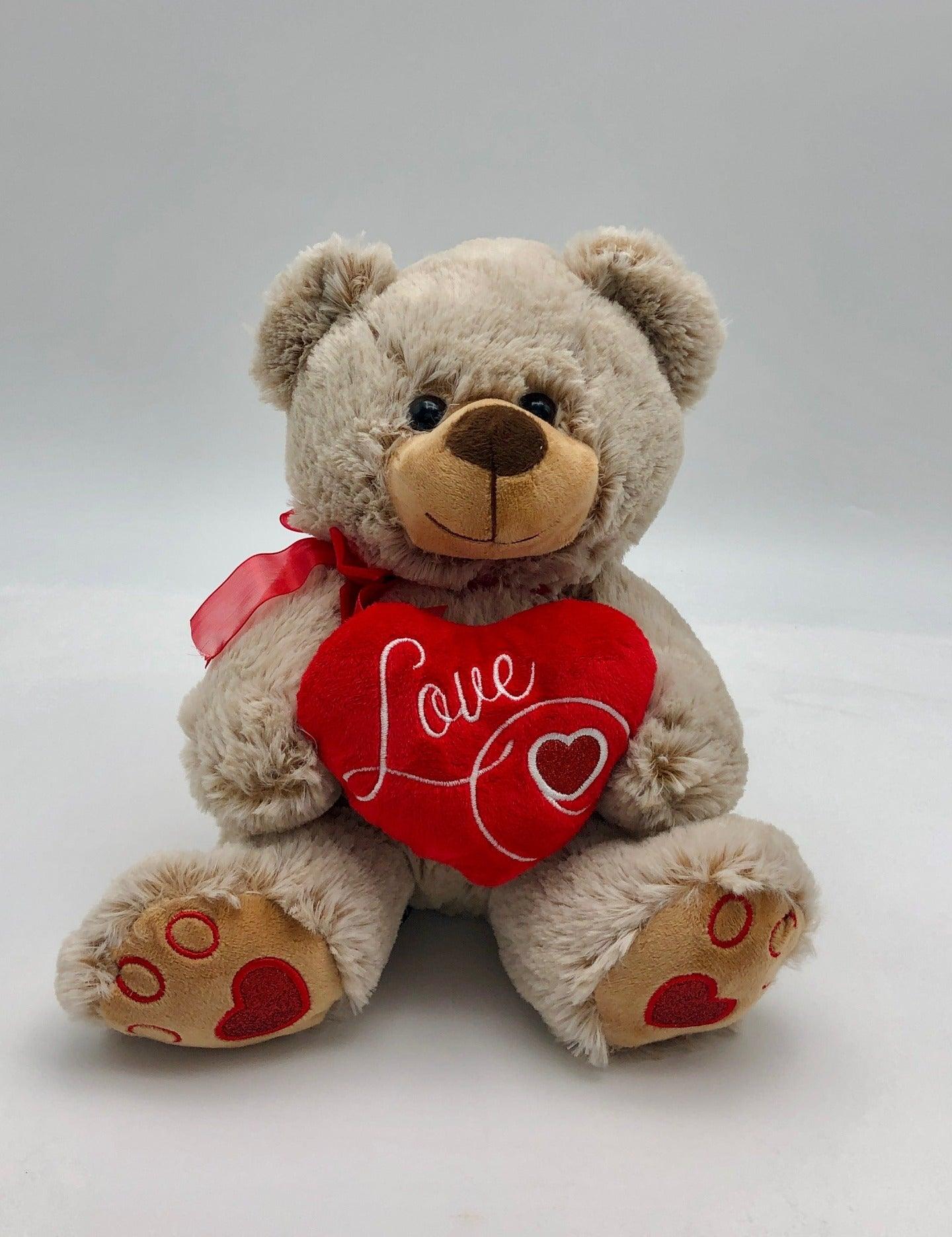 A photo of a charming light brown teddy bear. The teddy bear is adorned with a red ribbon around its neck and holds a red heart with the word âLoveâ written in white cursive letters.