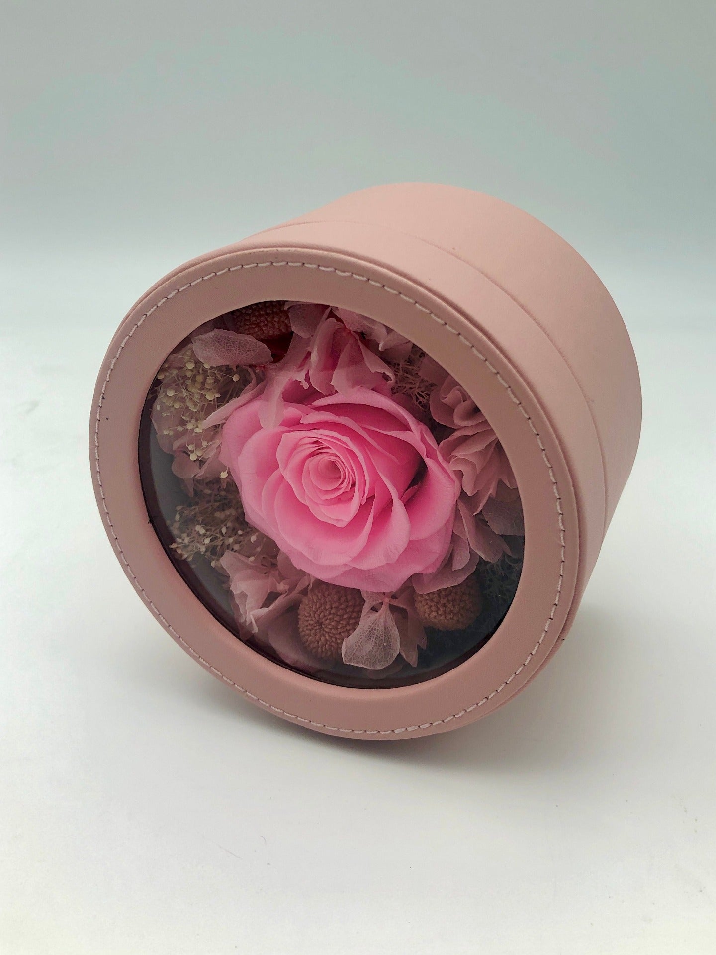A photo of a beautiful pink rose preserved in a round pink gift box. The rose is complemented by small, dried brown flowers, creating a charming and timeless gift perfect for any occasion.