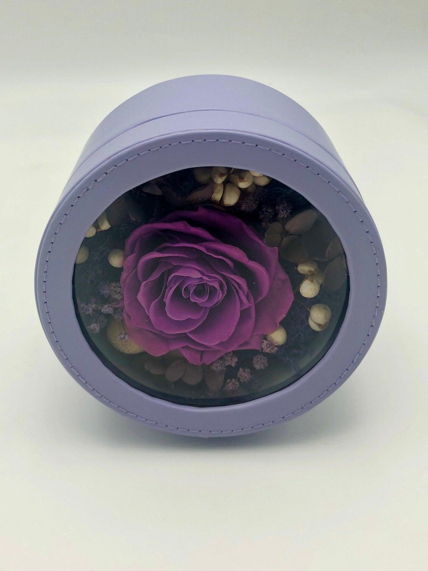 A photo of a stunning dark purple rose, beautifully preserved and displayed in a light purple round gift box. Itās a timeless symbol of elegance and beauty.