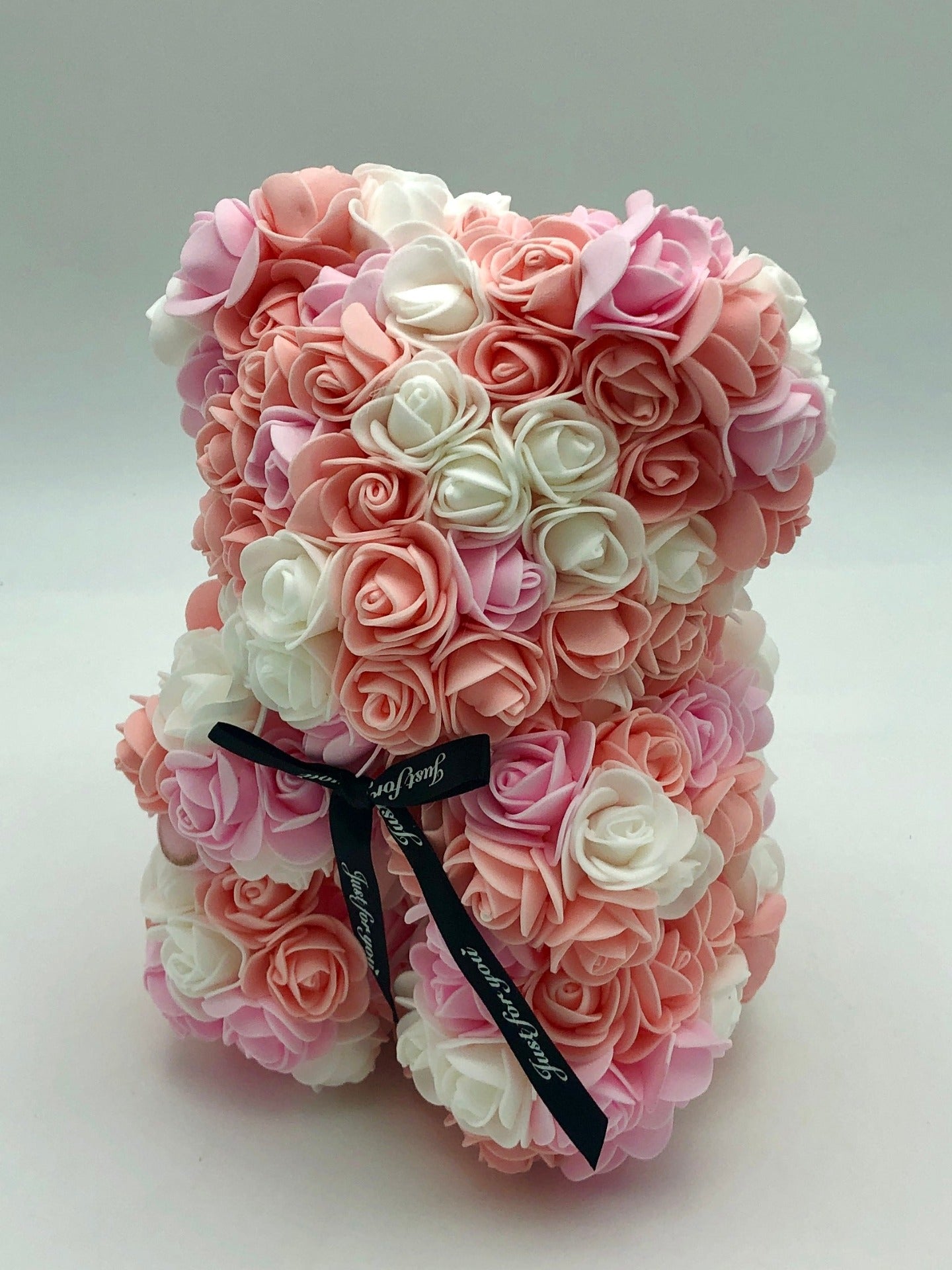 A teddy bear made from pink and white roses. The bear is standing upright and is adorned with a black ribbon. The background is plain white.