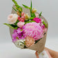 A dainty bouquet held against a neutral background, composed of blooming pink disbud mums, carnations, lisianthus, and assorted green leaves. The presentation is enhanced with rustic burlap wrapping and a branded Flowers Express Co tag. Made in Melbourne.