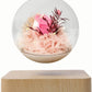 A glass snow globe with a wooden base, containing a bouquet of preserved flowers for delivery in Melbourne. The globe is lit from within with tiny fairy lights and the flowers are spilling out of the globe.