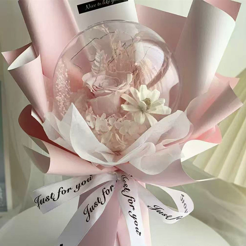A pink and white decorative flower bouquet with a clear round bubble containing dried flowers, wrapped in pink tissue paper and ribbon with printed text.