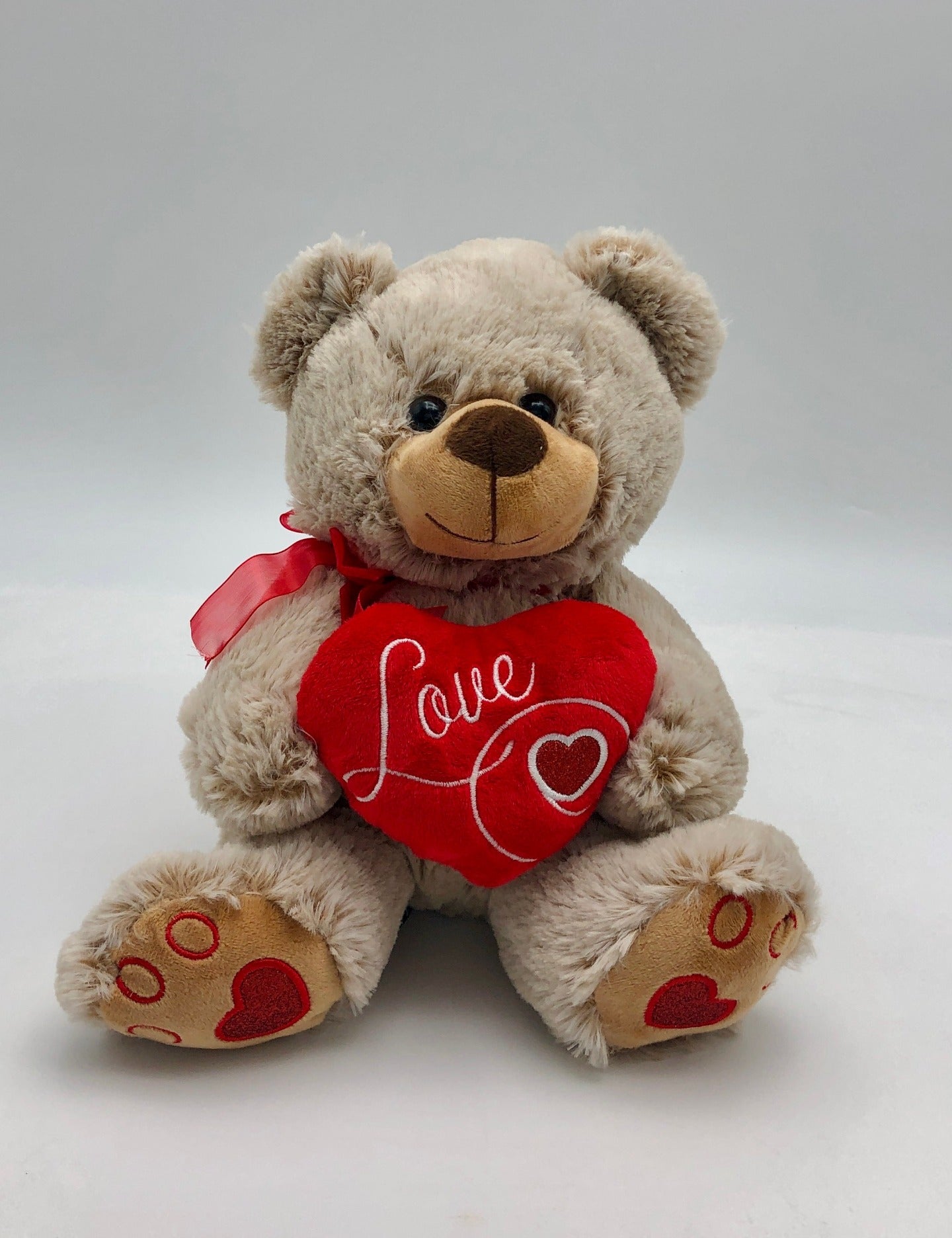 A photo of a charming light brown teddy bear. The teddy bear is adorned with a red ribbon around its neck and holds a red heart with the word “Love” written in white cursive letters.