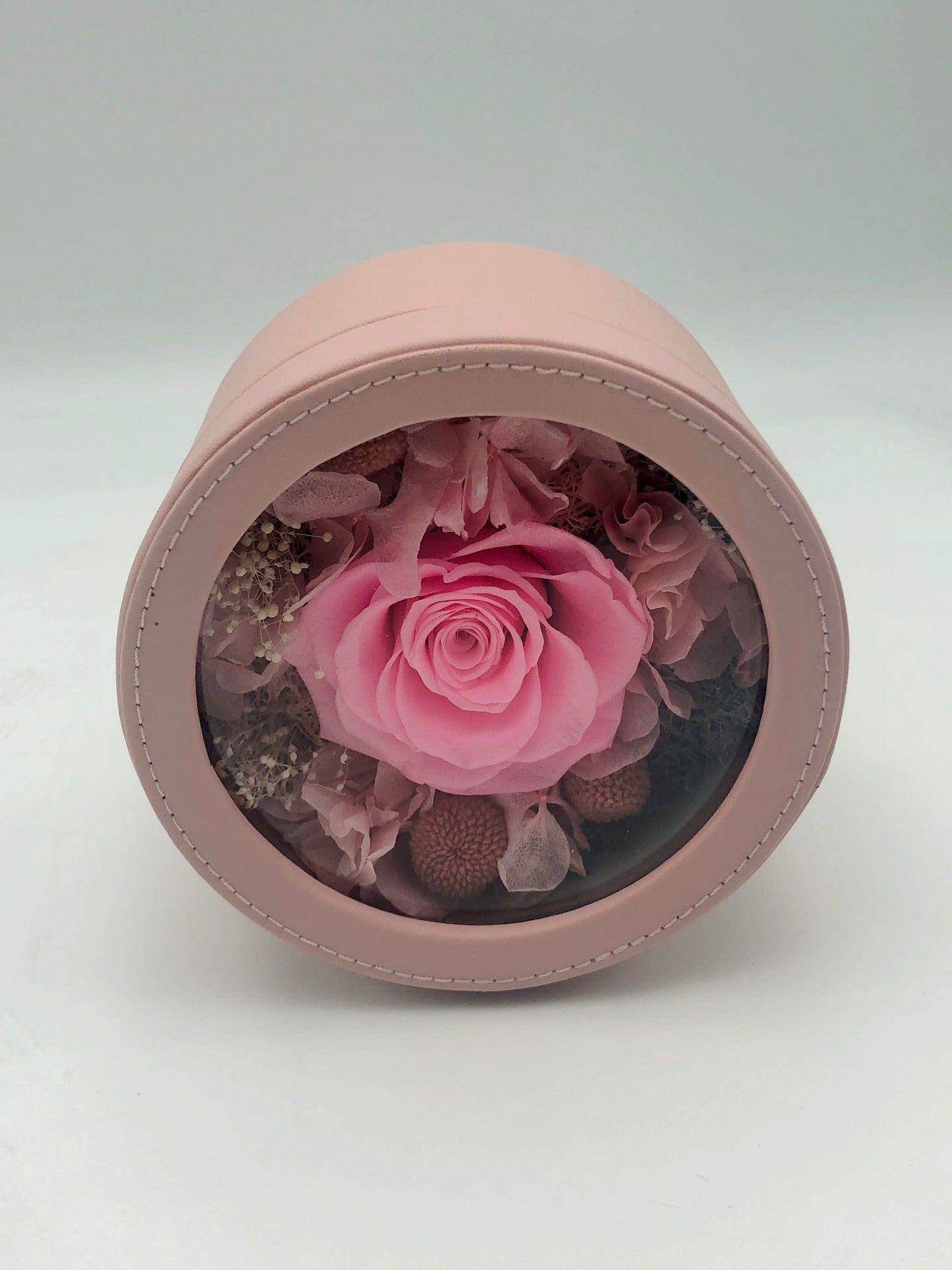 A photo of a lovely pink gift box containing a preserved pink rose and other flowers. The clear window on the lid allows a peek into the beautiful arrangement inside. Perfect for any special occasion!