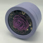A photo of a captivating purple rose, nestled among white and black flowers, beautifully presented in a light purple round gift box. A perfect symbol of love and elegance.