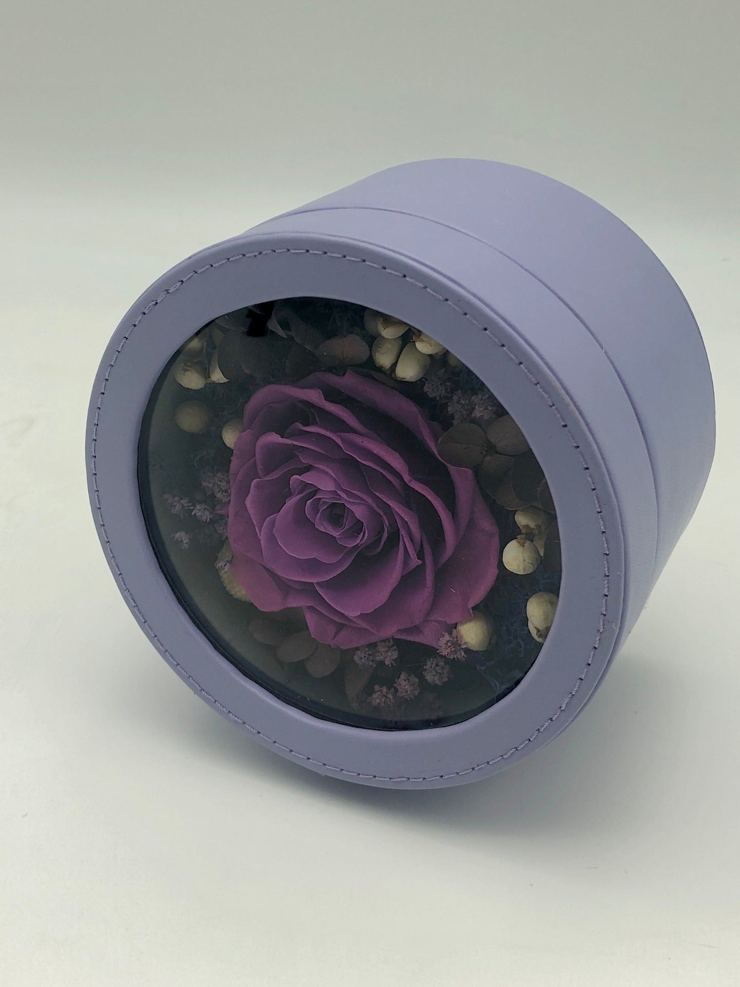 A photo of a captivating purple rose, nestled among white and black flowers, beautifully presented in a light purple round gift box. A perfect symbol of love and elegance.