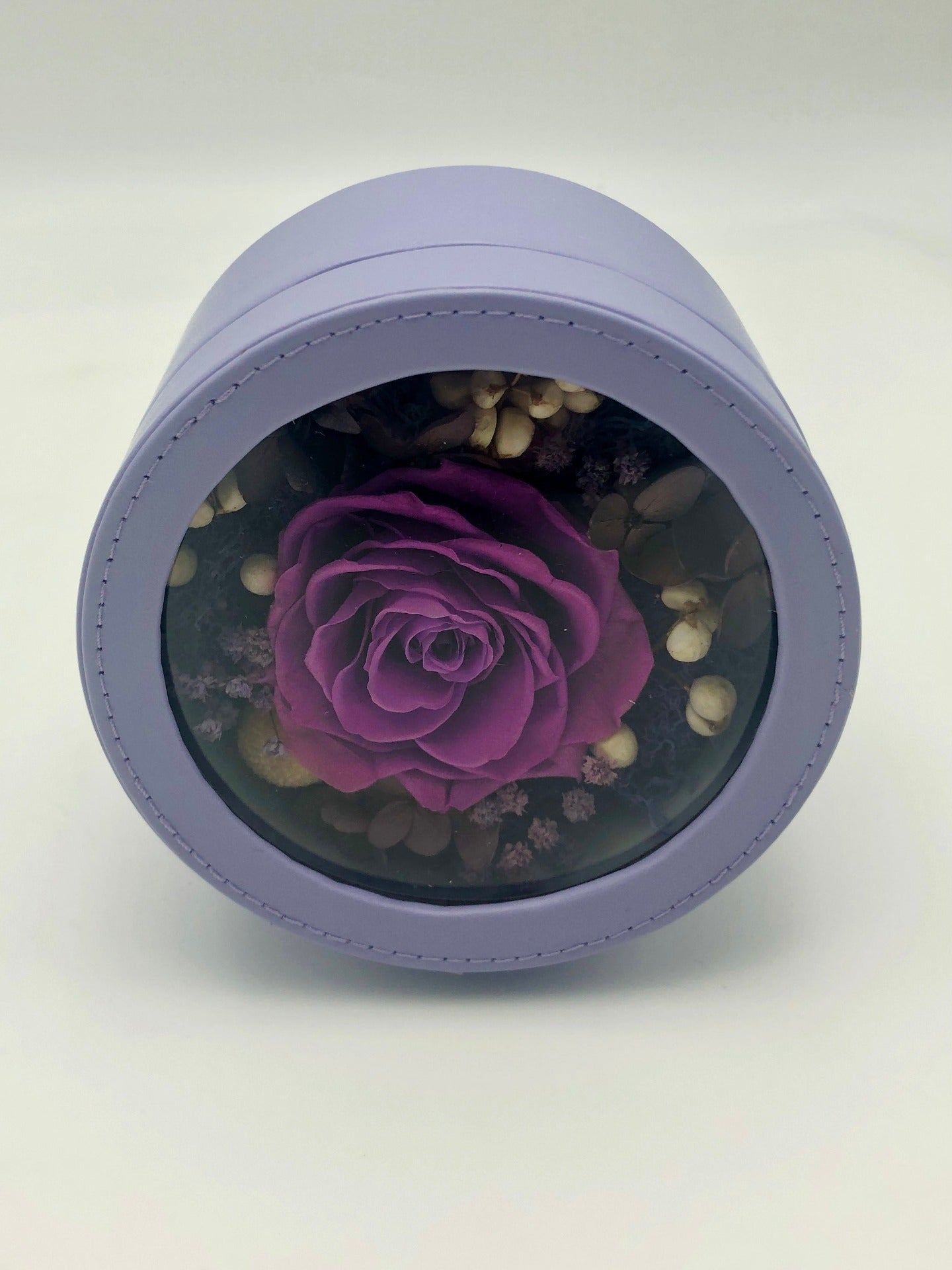 A photo of a stunning dark purple rose, beautifully preserved and displayed in a light purple round gift box. It’s a timeless symbol of elegance and beauty.