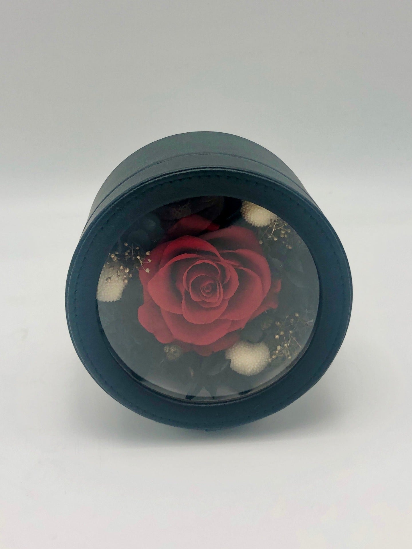 A photo of a preserved red rose, nestled among black and natural flowers in a black gift box. A perfect symbol of everlasting love and beauty.