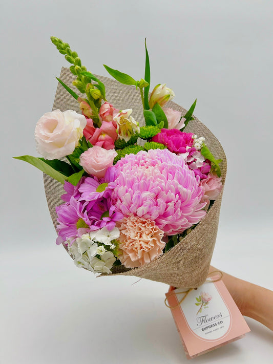 A dainty bouquet held against a neutral background, composed of blooming pink disbud mums, carnations, lisianthus, and assorted green leaves. The presentation is enhanced with rustic burlap wrapping and a branded Flowers Express Co tag. Made in Melbourne.