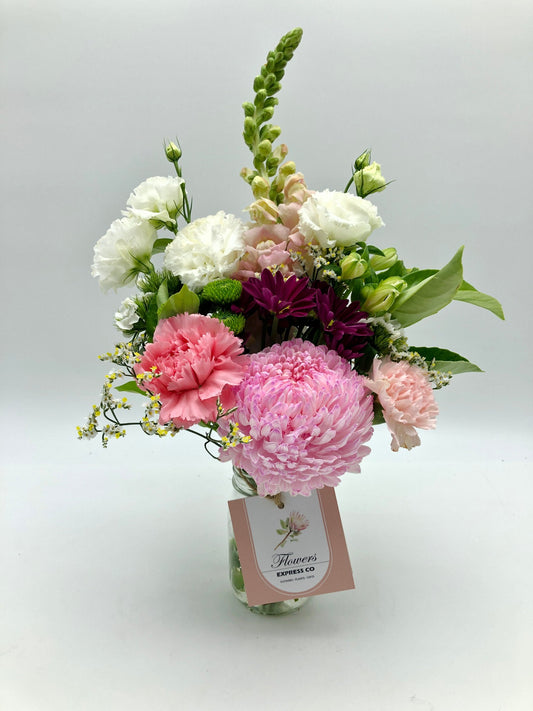 This is a photo of a beautiful flower bouquet in a vase. The vase is square-shaped and has a pink label that reads “Flowers Express Co.”. The bouquet comprises white, pink, and purple flowers with green leaves. The arrangement is such that the pink and purple flowers are in the centre, while the white flowers are on the outer edges. There are also small yellow flowers scattered throughout the bouquet. The background is plain white, making the flowers' vibrant colours stand out even more.