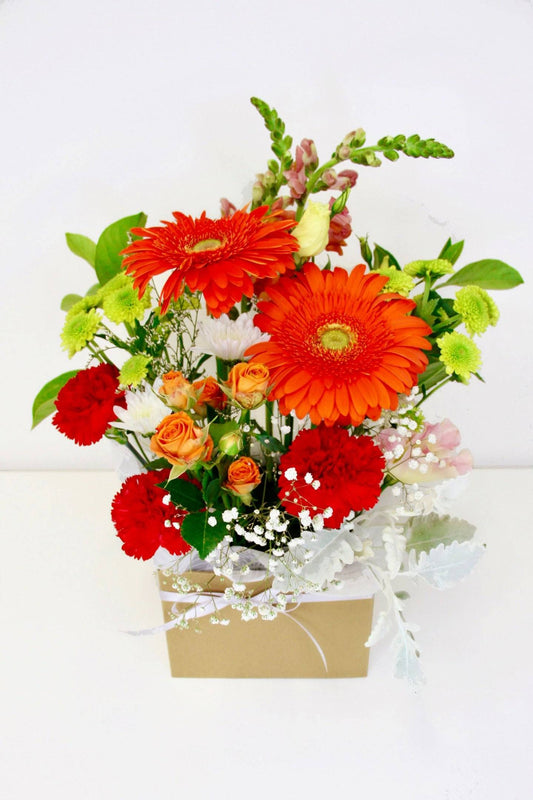 A flower arrangement of orange and red in a kraft box on a white table, excellent for gifting and decorating, available for delivery in Melbourne VIC.