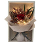 White box containing a red preserved rose, assorted dried flowers and a fairy light, encased in warm beige wrap and adorned with a grey ribbon.