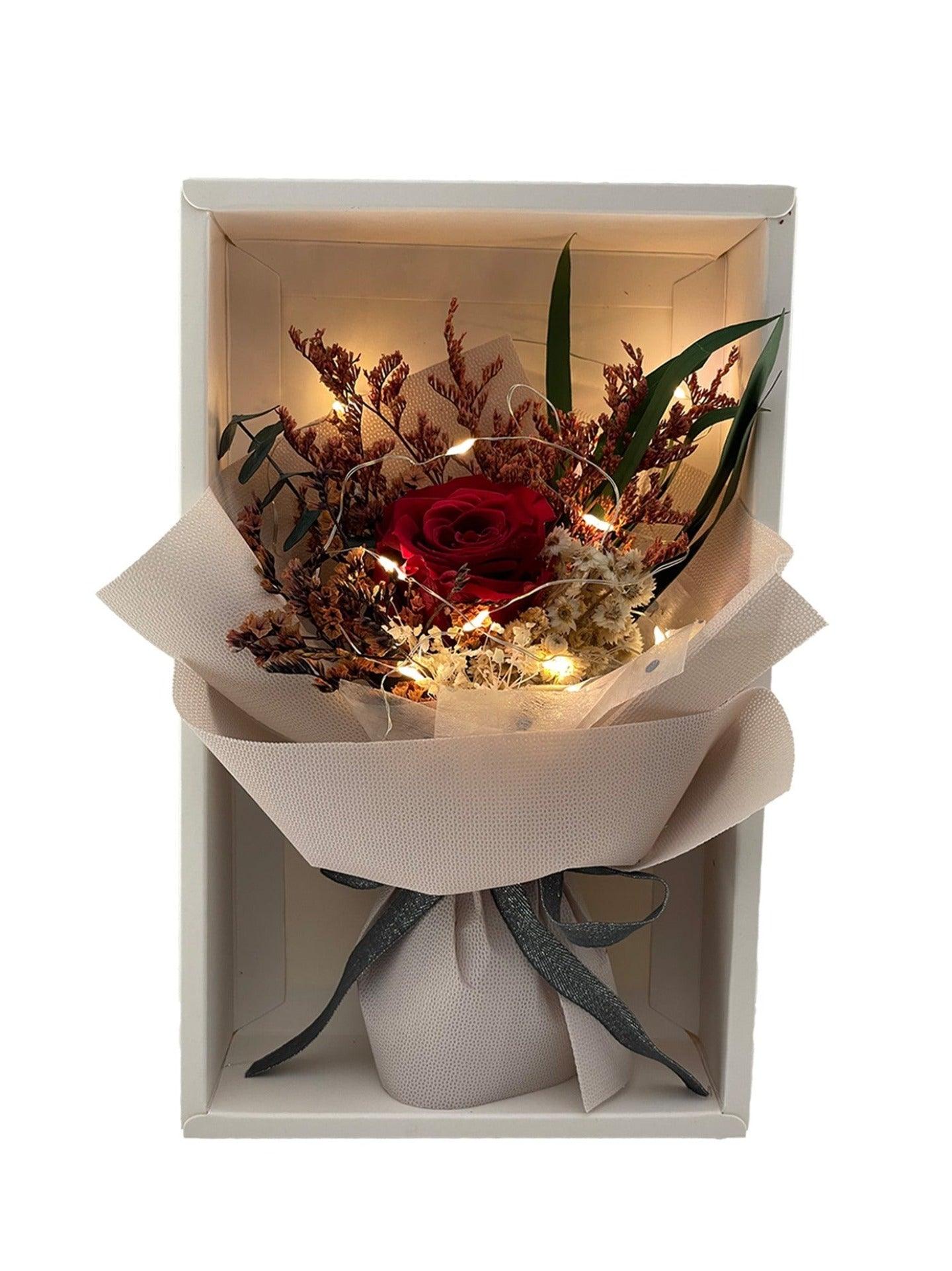 White box containing a red preserved rose, assorted dried flowers and a fairy light, encased in warm beige wrap and adorned with a grey ribbon.